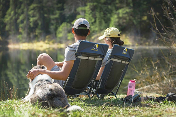Two people sitting in GC Outdoor Everywhere Chairs with a dog and a view of a pond